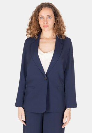 Donna con capelli ricci indossa una giacca blu navy sopra una maglietta bianca, in piedi davanti a uno sfondo chiaro e semplice, rivolta in avanti.