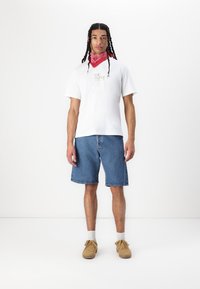 T-shirt en coton blanc avec texte doré, shorts en denim bleu, bandana rouge, baskets beiges et chaussettes blanches, se tenant devant un fond uni.