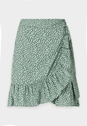 Saia de tecido verde com padrão de bolinhas pretas, com bainha franzida e design envolvente lateral. Textura suave e material leve.