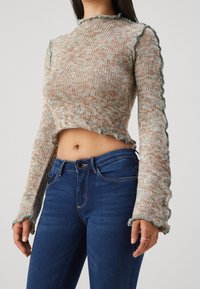 Suéter recortado de punto en beige moteado y azul con bordes festoneados, combinado con jeans ajustados azules. Tela texturizada y mangas largas.