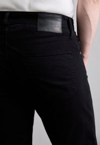 Fekete farmernadrág bőrből készült, gravírozott logóval ellátott folttal, öt zsebes kialakítással és diszkrét varrási részletekkel a hátsó zsebeknél.