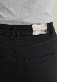 Mustad teksased, mille pind on sile, tagaküljel on taskud ja hõbedane silt, millel on kirjas "NEXT DENIM COLLECTION".