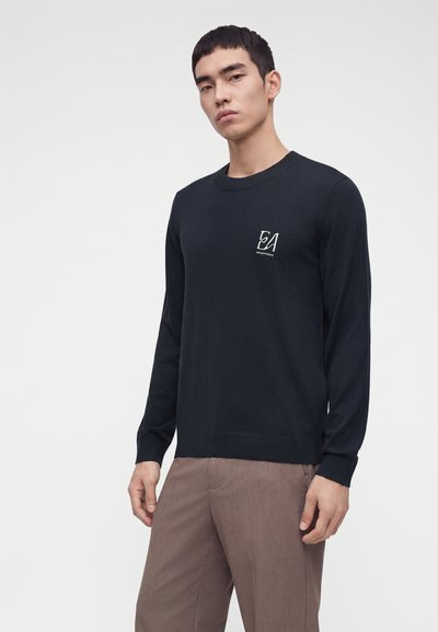 Jeune homme portant un pull noir à manches longues avec un petit logo blanc sur la poitrine et un pantalon marron, debout devant un fond uni.