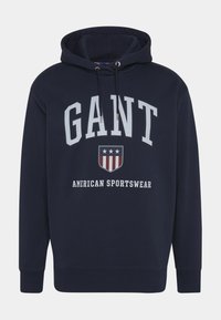 Navy blue huppari, joka on valmistettu puuvillasta, ja jossa on suuret valkoiset kirjaimet "GANT", shield-grafiikka täynnä tähtiä sekä teksti "AMERICAN SPORTSWEAR".