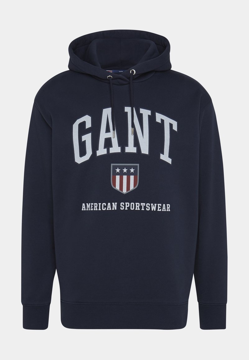 Navy blue huppari, joka on valmistettu puuvillasta, ja jossa on suuret valkoiset kirjaimet "GANT", shield-grafiikka täynnä tähtiä sekä teksti "AMERICAN SPORTSWEAR".