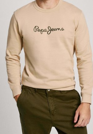 Beżowa bluza z długim rękawem z logo "Pepe Jeans", noszona z oliwkowozielonymi spodniami, ręce w kieszeniach, widoczny tułów, neutralne tło.
