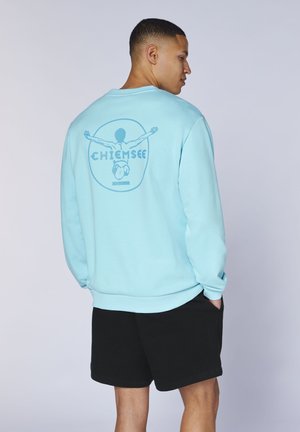 Mann trägt hellblauen Sweatshirt mit Grafik ausgestreckter Arme und Schriftzug „CHIEMSEE“ auf dem Rücken, kombiniert mit schwarzen Shorts, Hände in den Taschen.