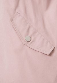 Ljust rosa tyg med en slät textur, med en lockficka och en silverfärgad tryckknapp. Sömmarna är synliga längs fickans kanter.