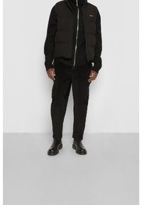 Schwarze Steppweste über einem schwarzen gerippten Pullover, kombiniert mit schwarzen Kord-Hosentaschen und schwarzen Leder-Stiefeletten.