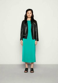 Mulher vestindo um vestido midi teal, jaqueta de couro preta, sandálias abertas pretas, em pé contra um fundo claro e liso.