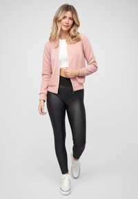 Světle růžová prošívaná bomber bunda s předním zipem, v kombinaci s černými legínami a bílými teniskami. Jednoduchý design a hladká textura.