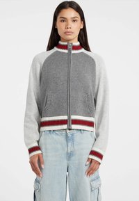 Maglione corto zinco grigio e grigio chiaro con dettagli a coste rossi e bianchi, tasche laterali e colletto alto in un materiale morbido.