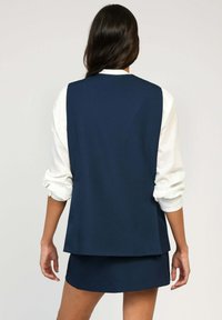 Gilet sans manches bleu marine avec une coupe décontractée par-dessus une chemise blanche à manches longues. Tissu lisse, design simple et détails minimalistes.