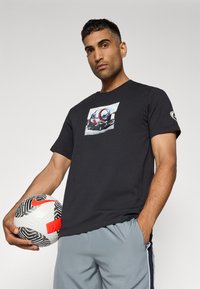 Sort bomulds t-shirt med et grafisk print af et hjelme og en racerbils. Modellen holder en hvid og sort fodbold med røde accenter.