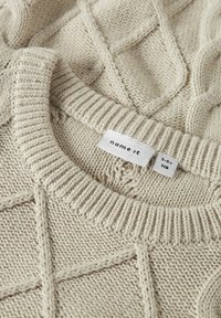 Maglione beige lavorato a maglia con un motivo a diamante testurizzato, collo a giro ribattuto e un'etichetta che mostra marca e taglia. Materiale morbido e di peso medio.