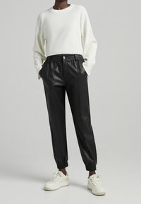 Mustat keinonahkaiset jogger-housut, joissa on joustava vyötärönauha ja resorit nilkoissa, yhdistetty valkoiseen ribbineulottuun puseroon ja valkoisiin tennareihin.