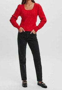 Roter gestrickter Pullover mit einem quadratischen Ausschnitt und strukturiertem Muster, kombiniert mit schwarzen High-Waist-Jeans und schwarzen Slippern.