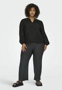 Top nero a maniche lunghe con scollo a V e polsini elasticizzati, abbinato a pantaloni larghi grigio scuro a trama e sandali neri con suola spessa.