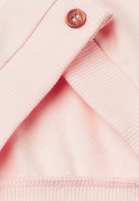 Světle růžový límec mikiny s žebrovanou texturou, vybavený jedním hnědým knoflíkem a měkkou, hladkou tkaninou. Viditelný detail šití.