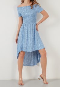 Světle modré šaty s odkrytými rameny, krátkými rukávy, nařaseným pasem a asymetrickým spodním okrajem, který má bílý květinový vzor. Průhledné podpatky.