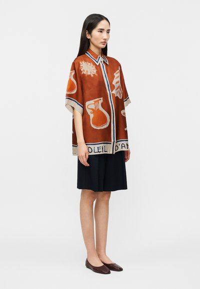 Γυναίκα που στέκεται φορώντας ένα φαρδύ πουκάμισο σε σκουρό πορτοκαλί χρώμα με αφηρημένα άσπρα και μπλε μοτίβα και μαύρη φούστα μέχρι το γόνατο με καφέ φλατ παπούτσια.