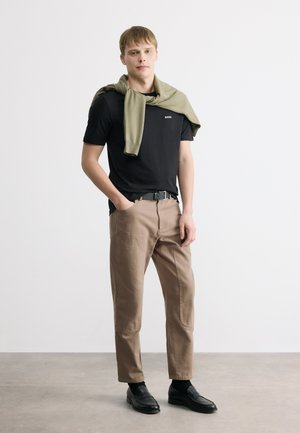 Mladý muž stojaci s rukami v džepoch, oblečený v čiernom tričku, svetlohnedých nohaviciach, čiernych topánkach a zelenom svetri prehodenom cez plecia.