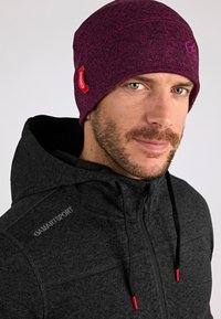Gorro morado con un patrón de punto texturizado, que presenta una etiqueta de acento roja. Lleva puesto un sudadera negra con cremallera, que exhibe una textura de tela suave.