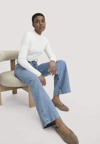 Valkoinen pitkähihainen paita, siniset korkeavyötäröiset leveälahkeiset farkut ja ruskeat ballerinat. Aihe istuu vaaleasta puusta valmistetulla tuolilla, jossa on beige tyyny.