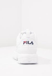 Zapatilla blanca con parte superior sintética texturizada, cuello acolchado y una suela de goma flexible. Presenta el logo de Fila en rojo, azul y blanco.
