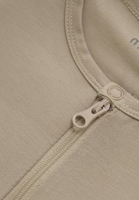 Beige plagg med en slät textur, som har en framtill placerad dragkedja och ribbad krage. Dragkedjens dragningsdel är av en ljus färg som smälter in med tyget.
