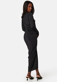 Długa czarna sukienka z długimi rękawami, marszczonymi detalami po bokach, smukłym krojem i długością maxi. Połączona z czarnymi sandałami na obcasie.