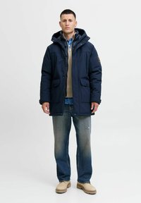Mørkeblå pufferjakke med hætte, med frontlommer og lynlås. Bæres over en beige strikket sweater, parret med slidte blå jeans og brune støvler.