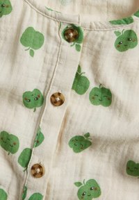 Leichter Stoff mit einem cremfarbenen Hintergrund, der kleine grüne Apfelmuster und braune Knöpfe entlang der Vorderblende zeigt. Strukturierte Oberfläche.