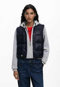 Chaleco acolchado azul marino con diseño de rombos, cierre delantero a presión y dobladillo con cordón, usado sobre una sudadera gris y combinado con vaqueros azules.