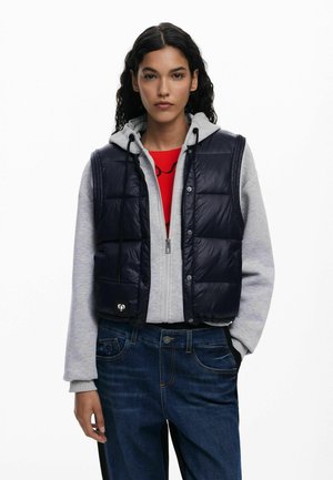 Navy puffer vest with quilted design, front snap closure, and drawstring hem, worn over a gray hoodie and paired with blue jeans.