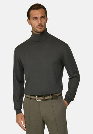 Homme portant un pull à col roulé gris foncé et un pantalon marron avec une ceinture tressée, debout devant un fond clair uni.