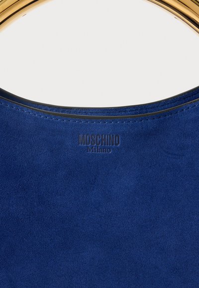 Kék velúr kézitáska arany kör alakú fogantyúval és a "MOSCHINO Milano" logóval, amely az alsó szélén van bélyegezve.