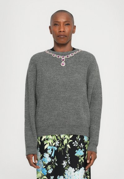 Pull gris à côtes avec un encolure ornée de bijoux, présentant des accents roses et transparents. Porté par-dessus une jupe à motifs floraux aux tons bleus et verts.