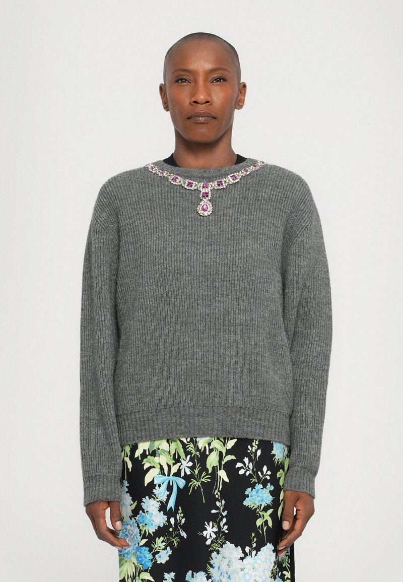 Grauer, gerippter Pullover mit einem mit Schmucksteinen besetzten Halsausschnitt, der rosa und klare Akzente setzt. Getragen über einem rock mit floralem Muster in Blau- und Grüntönen.