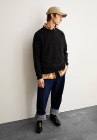 Schwarzer Rippenpullover, orange-kariertes Hemd darunter, blaue weite Jeans mit hochgekrempelten Säumen, schwarze Slipper und eine beige Kappe.