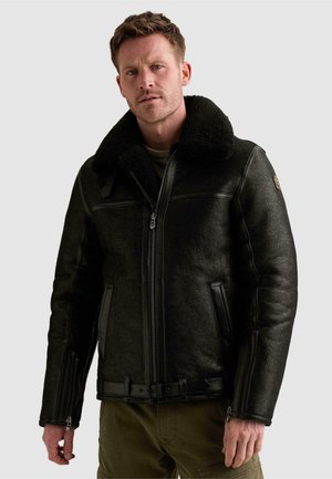 Homme portant une veste en cuir d’agneau noire avec grand col, fermeture éclair devant et taille bouclée, debout devant un fond gris clair.