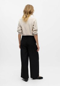 Donna con capelli biondi che indossa un maglione beige, pantaloni neri a gamba larga e scarpe nere, in piedi di spalle contro uno sfondo bianco uniforme.