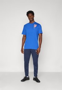 Blå sports t-shirt med rund halsudskæring og logo på brystet, parret med marineblå træningsbukser og sorte sportsko.