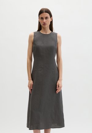 Femme aux cheveux bruns raides portant une robe ajustée sans manches, gris foncé, longueur cheville, sur un fond clair uni.