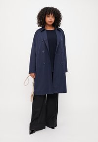 Marinblå dubbelknäppt trenchcoat med avslappnad passform, säkrad med knappar, i kombination med svarta vida byxor och en beige handväska.