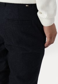 Marinblå byxor i fiskbensmönster med bakficka. Bärs med en vit ribbad tröja, vilket ger en avslappnad passform och rak bendesign.