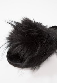 Mule noire avec une tige en fausse fourrure noire pelucheuse. La semelle est plate et l'intérieur est rembourré pour plus de confort.