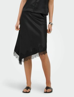 Femme portant une jupe en satin noire avec un ourlet en dentelle asymétrique, un haut noir sans manches, des sandales noires, un bracelet en argent et une bague sur fond blanc.