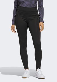 Zwarte high-waisted leggings van een gladde, rekbare stof, met zijnaad en een platte tailleband, gecombineerd met witte sportschoenen.