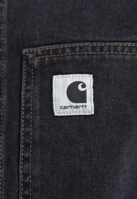 Mustast denimist koosneb valgest ruudukujulisest logopatchist, millel on must stiliseeritud "C" ja "carhartt" tekst. Näha on õmbluse detaili taskul.
